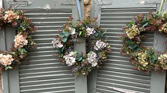 Three rustic floral window shutters with faux flower wreaths, set against a stone wall background.