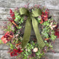 An autumn-themed artificial wreath with green and burgundy eucalyptus leaves, red berries, cinnamon sticks, and a large olive green ribbon bow, displayed on a weathered gray wooden wall.