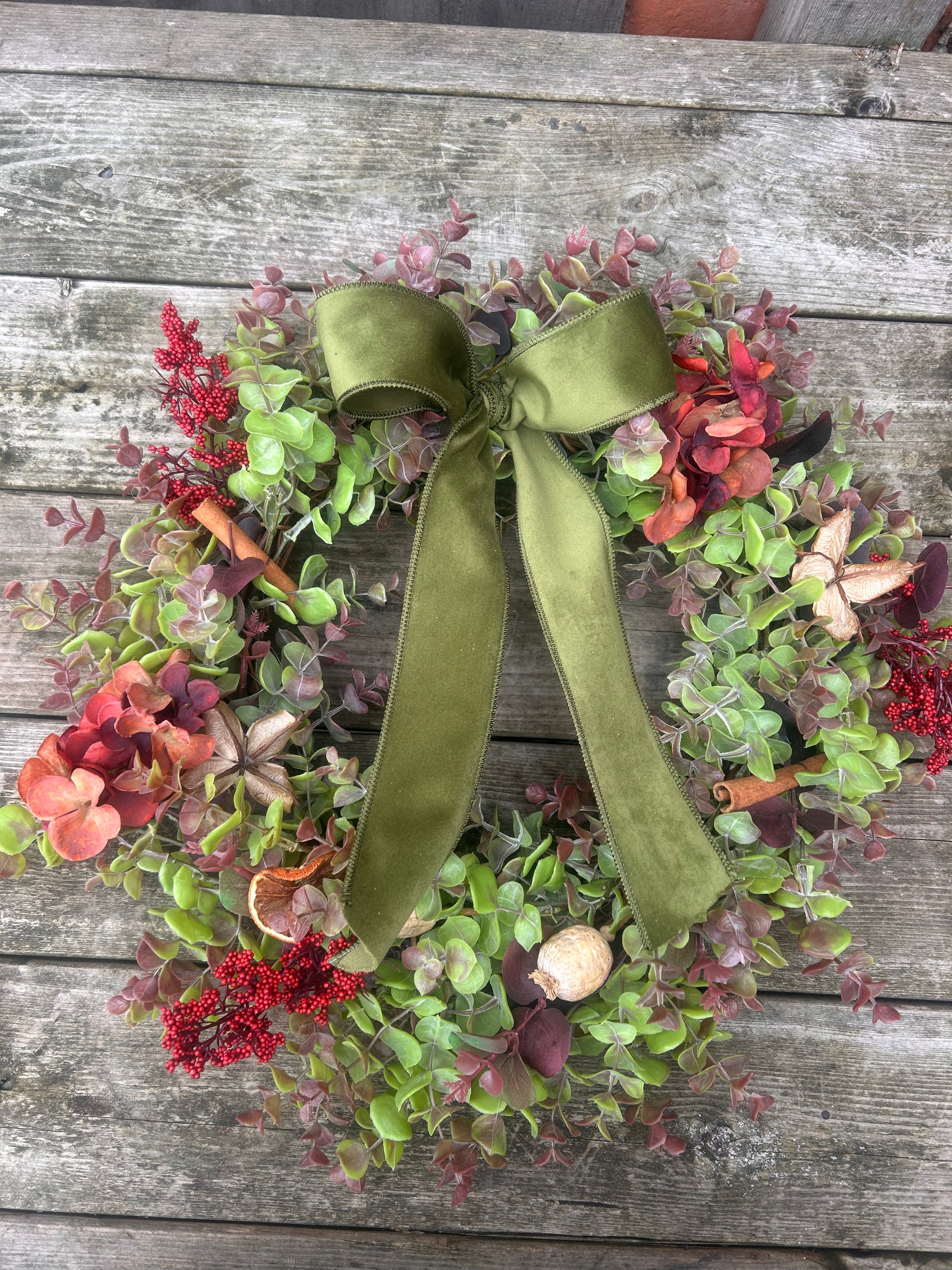 An autumn-themed artificial wreath with green and burgundy eucalyptus leaves, red berries, cinnamon sticks, and a large olive green ribbon bow, displayed on a weathered gray wooden wall.