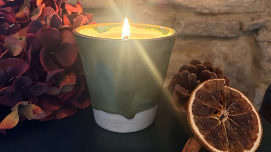 A lit candle in a ceramic holder sits on a dark surface against a rustic stone wall, styled with dried orange slices, a cinnamon stick, pinecones, and red flowers.