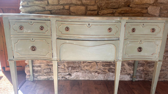 A distressed French-style console table with a sage green painted finish and gold leaf detailing, set against a rustic stone wall and dark wood floor.