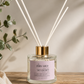 A clear glass reed diffuser bottle with a pale pink label reading 'The BARN SHED' and 'MULBERRY,' topped with a gold cap and white reeds, set on a wooden surface with soft shadows and greenery in the background.