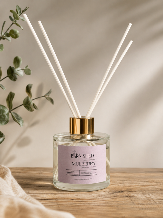A clear glass reed diffuser bottle with a pale pink label reading 'The BARN SHED' and 'MULBERRY,' topped with a gold cap and white reeds, set on a wooden surface with soft shadows and greenery in the background.
